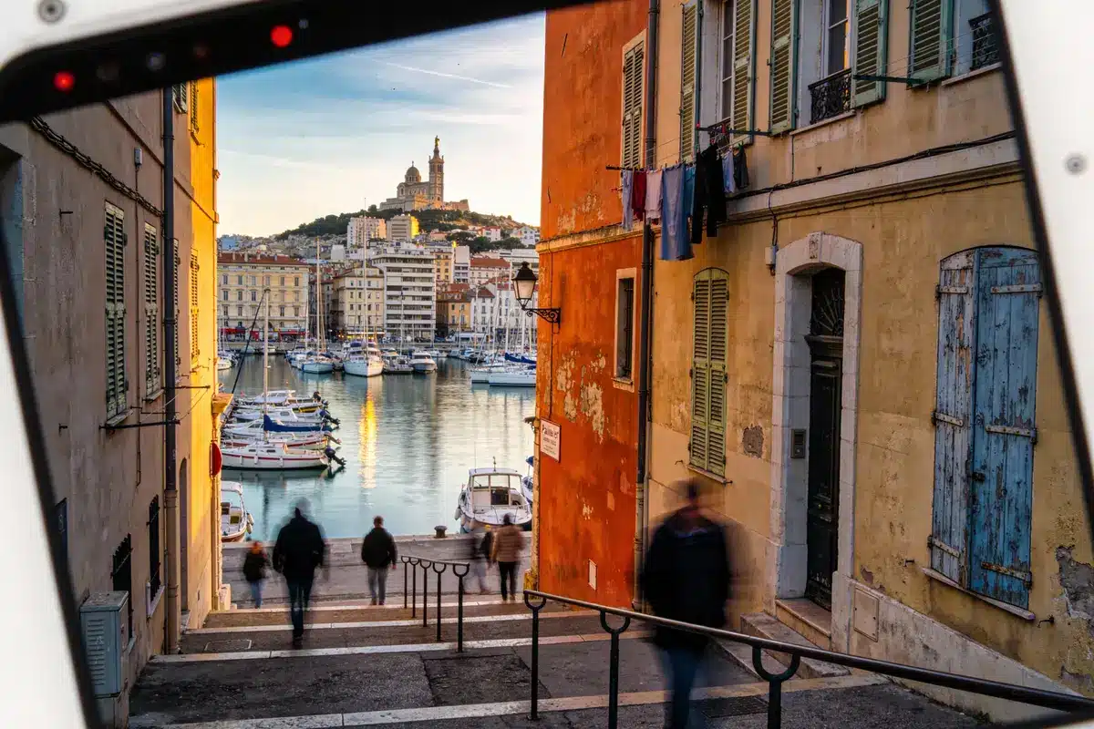 Ruelle avec vue sur le port de Marseille, bateaux, basilique Notre-Dame de la Garde en arrière-plan, personnes floues.