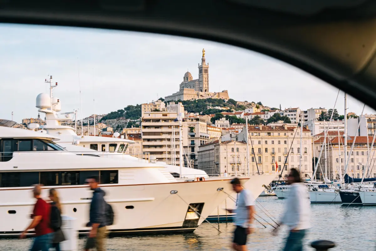 Vue depuis un véhicule sur un port animé avec yachts, bâtiments et colline en arrière-plan.