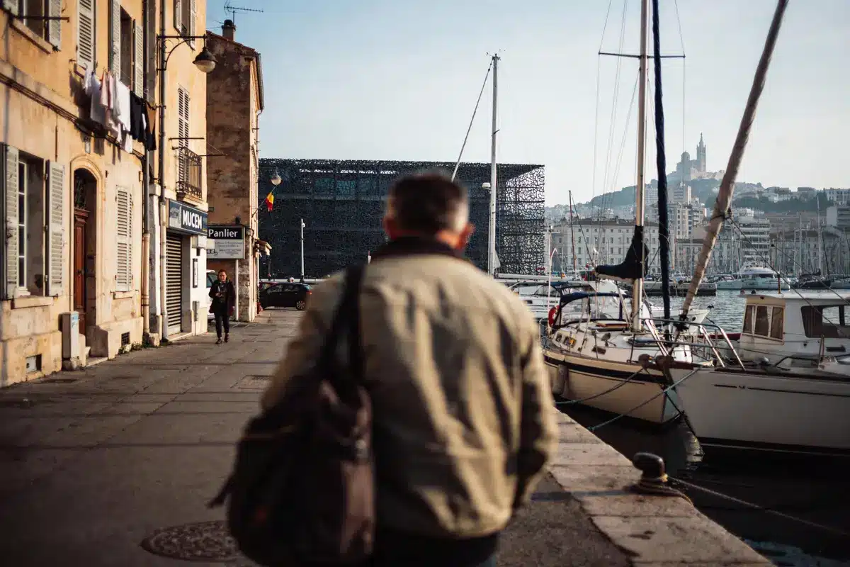 Un homme marche sur un quai bordé de bateaux, bâtiments historiques en arrière-plan.