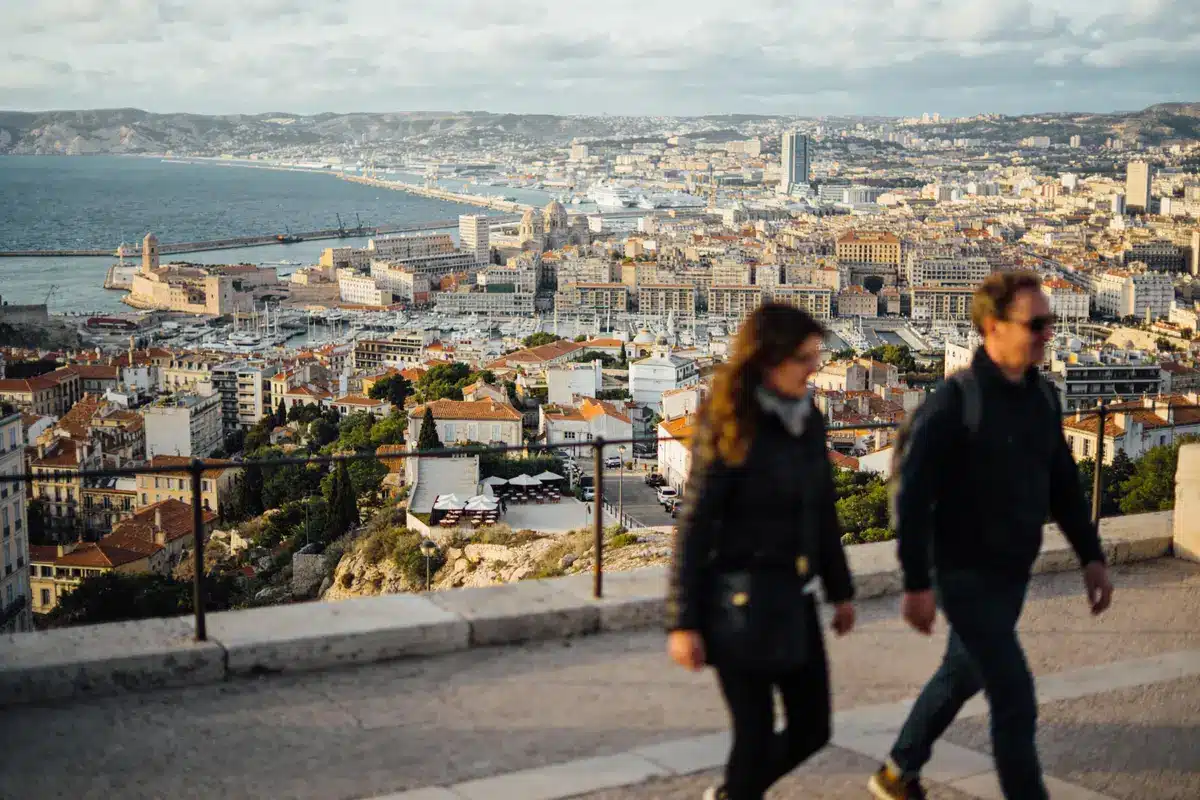 Vue panoramique de Marseille avec mer, bâtiments, fort, personnes floues marchant au premier plan, ciel nuageux.