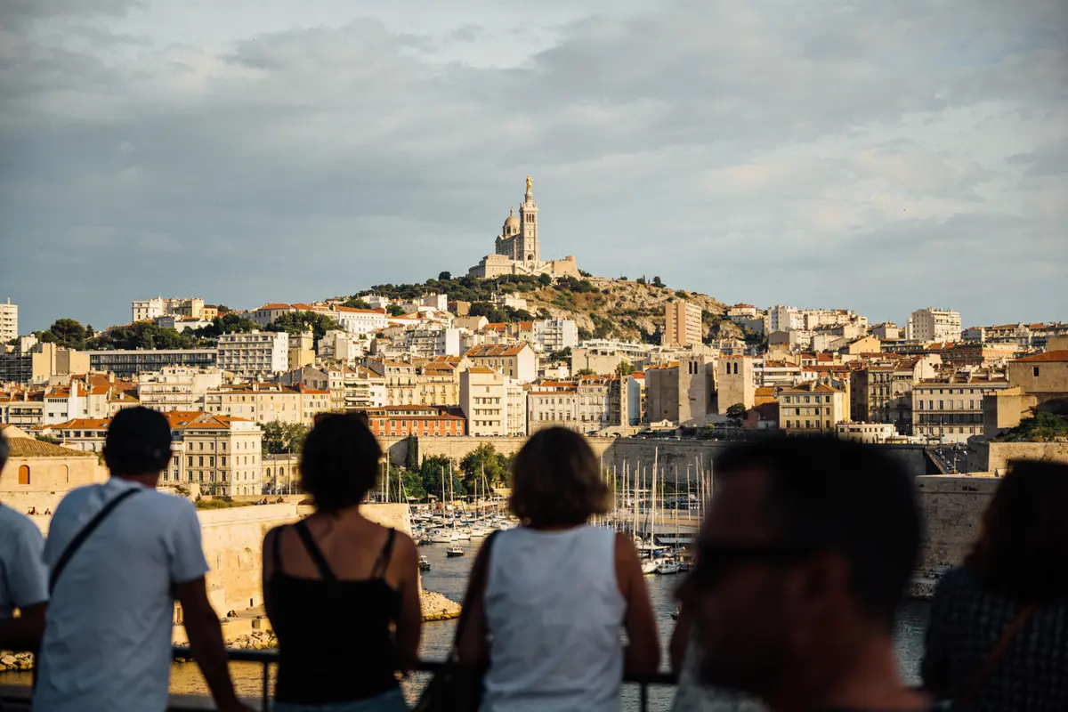 Des gens regardent la ville de Marseille, avec la basilique Notre-Dame de la Garde sur une colline.