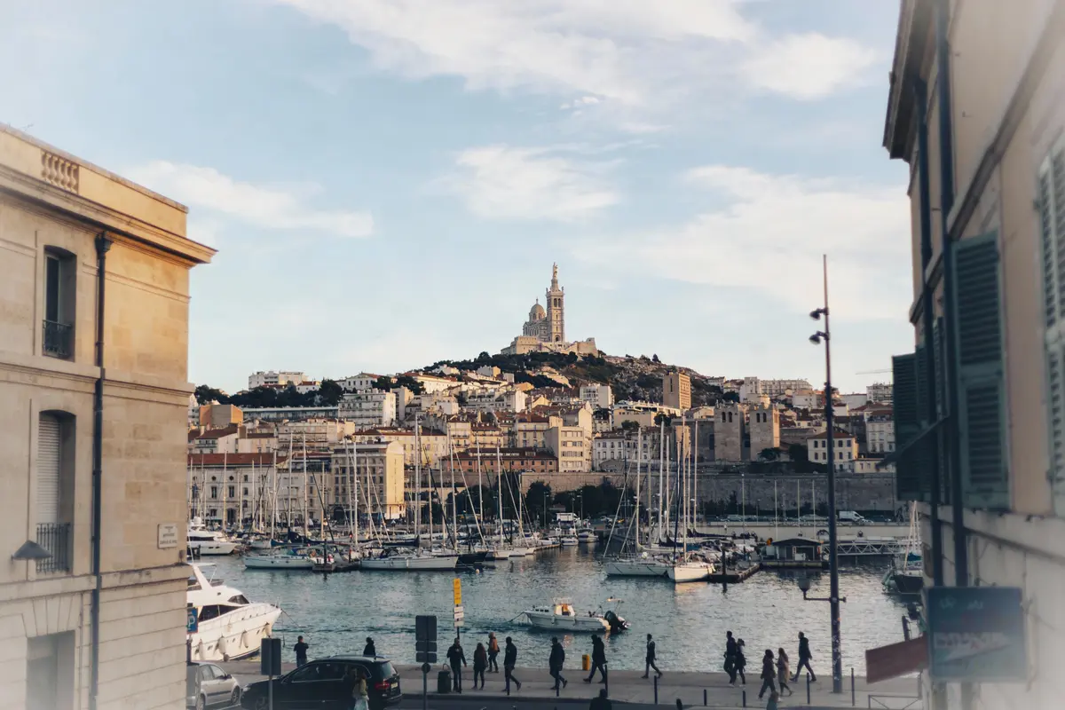 Vue de Marseille avec port de plaisance, basilique Notre-Dame de la Garde, bâtiments historiques et ciel partiellement nuageux.