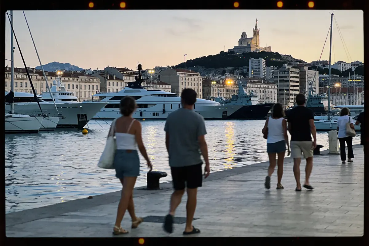 Personnes marchant le long du quai avec vue sur des yachts et une église sur la colline au crépuscule.