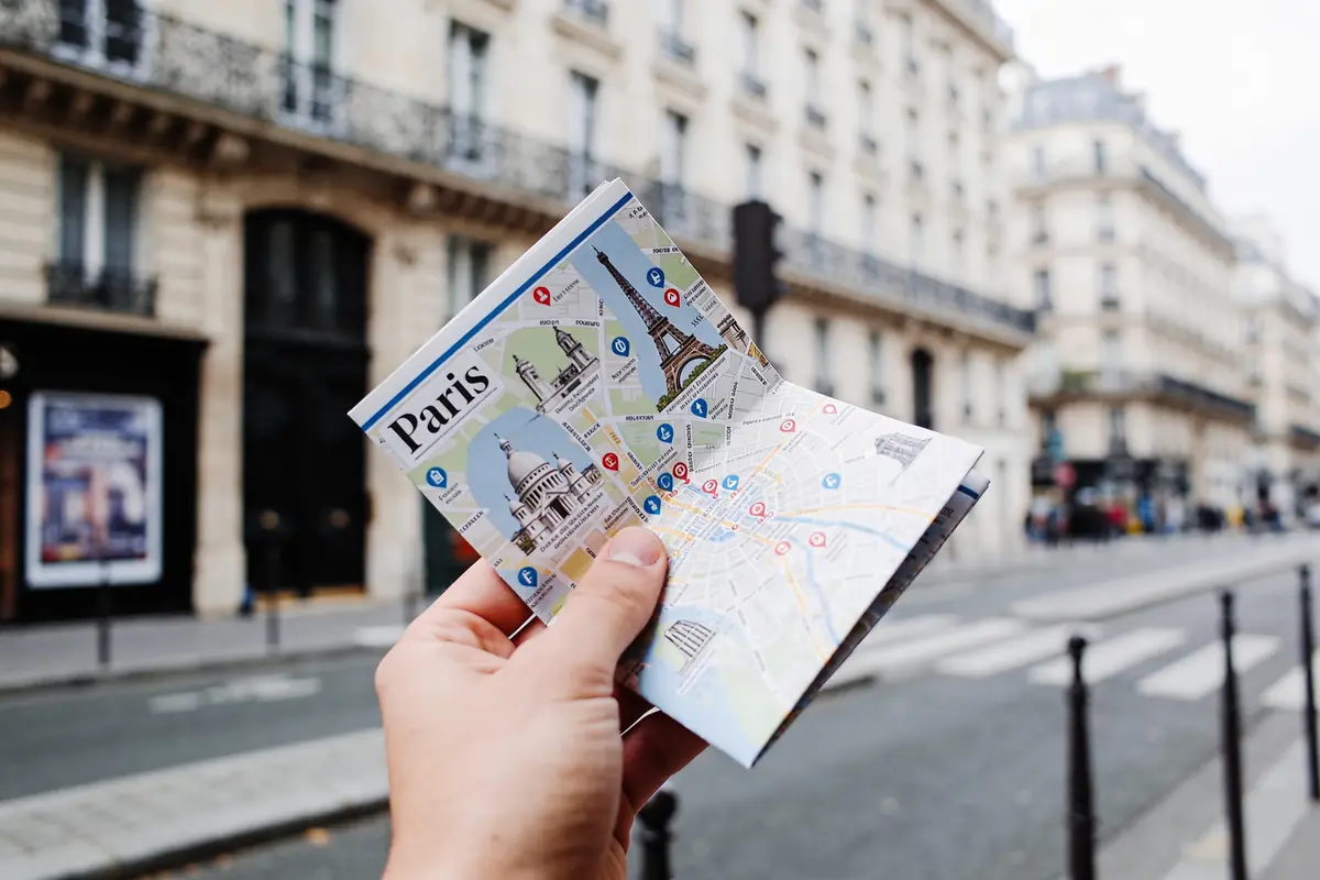 Une main tient une carte touristique de Paris, rue bordée d'immeubles haussmanniens en arrière-plan.