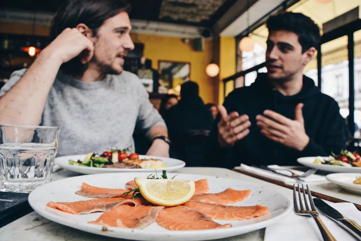 Deux personnes discutent dans un restaurant, assiettes de saumon fumé et salades sur la table, ambiance conviviale.