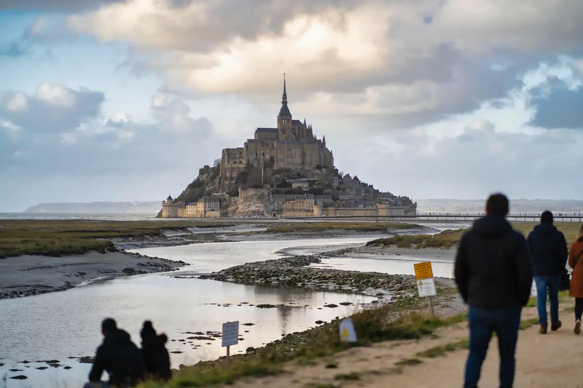 Paysage du Mont-Saint-Michel avec visiteurs au premier plan, ciel nuageux et rivière sinueuse.