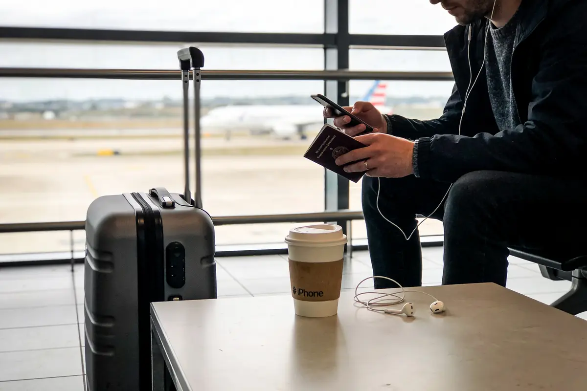 Homme à l'aéroport avec valise, téléphone, passeport, café à emporter, écouteurs et avion en arrière-plan.