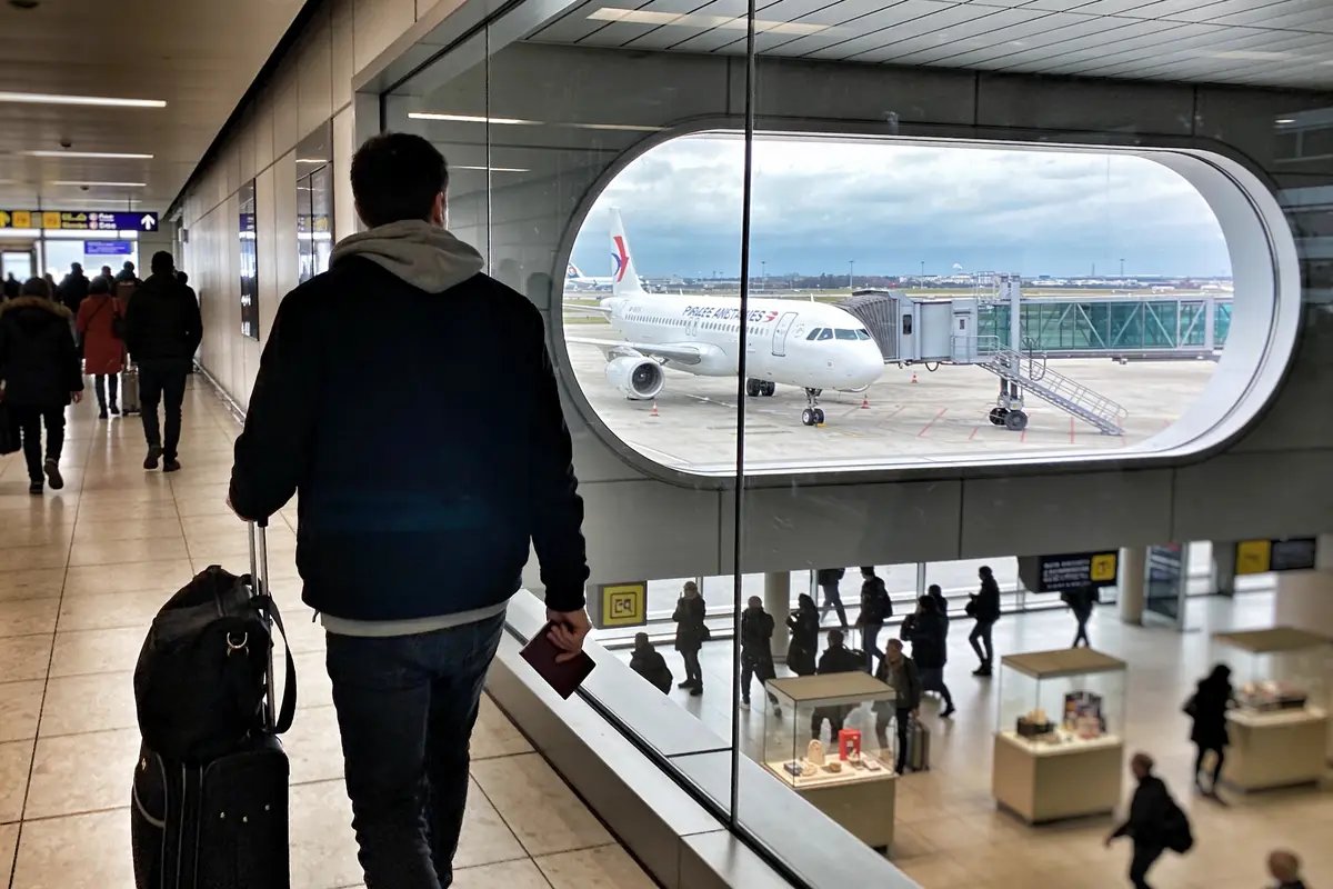 Personne avec valise et passeport, regarde un avion sur le tarmac à travers une grande fenêtre, aéroport occupé.