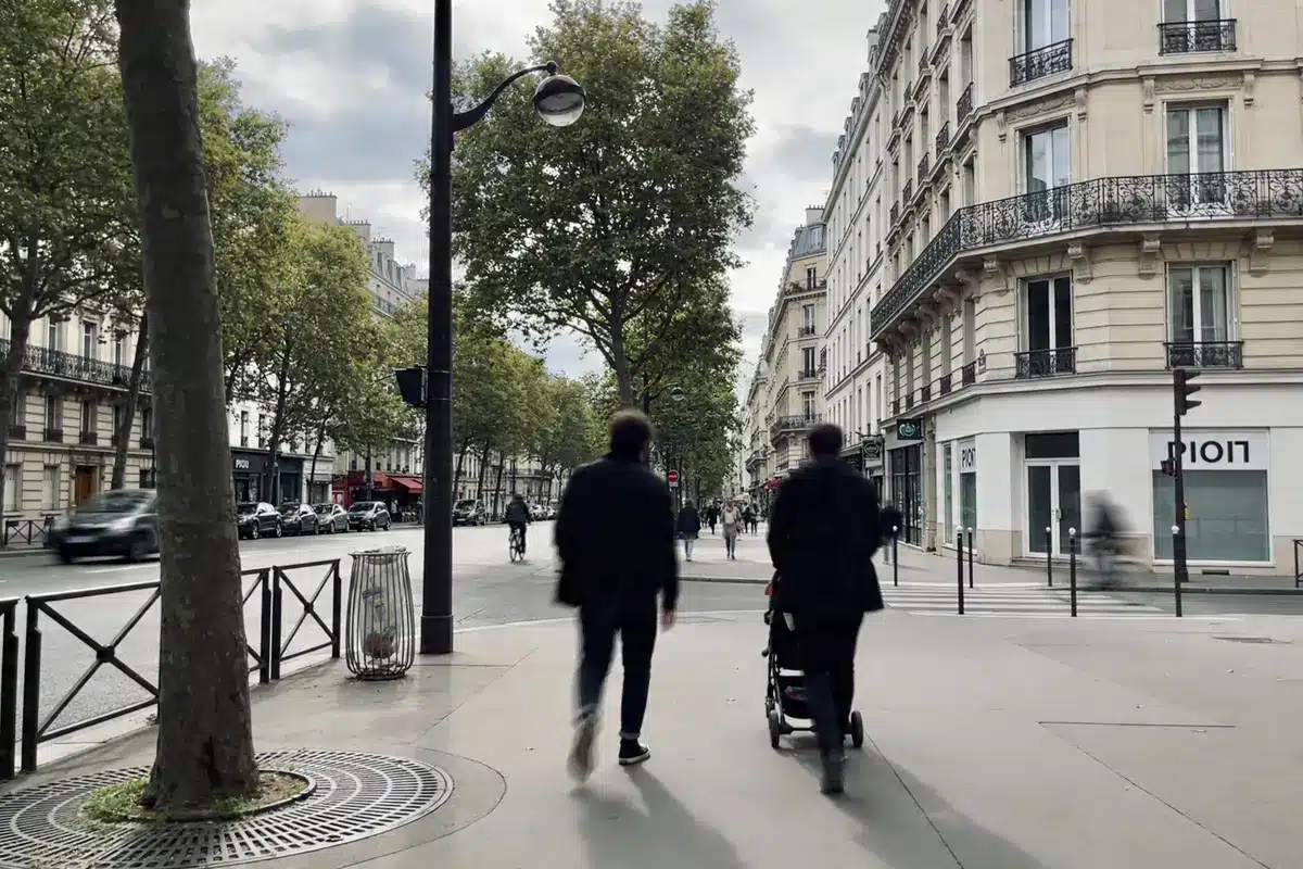 Deux personnes marchent sur un trottoir parisien bordé d'immeubles haussmanniens et d'arbres, ciel nuageux.