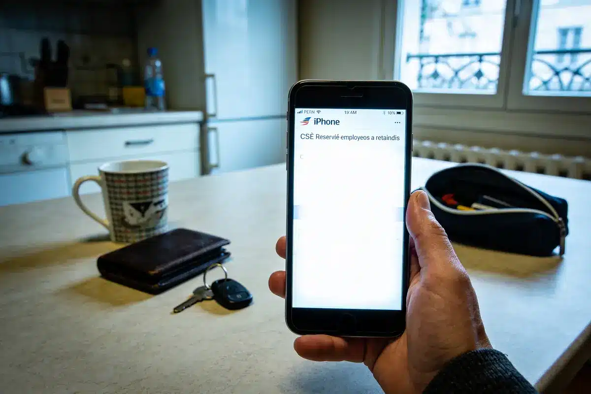 Homme tenant smartphone affichant "CSE Réservé employés", avec tasse, portefeuille et clés sur la table de cuisine.