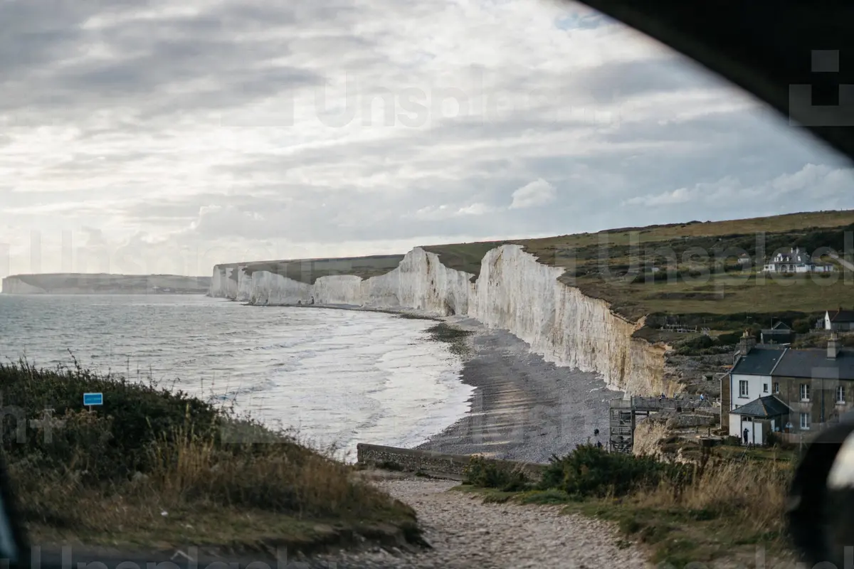 Falaises blanches surplombant la mer, maisons et sentier herbeux au premier plan, ciel nuageux.