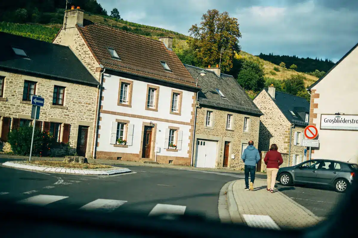Un village avec maisons en pierre, colline, deux personnes, panneau de rue, et voiture stationnée.