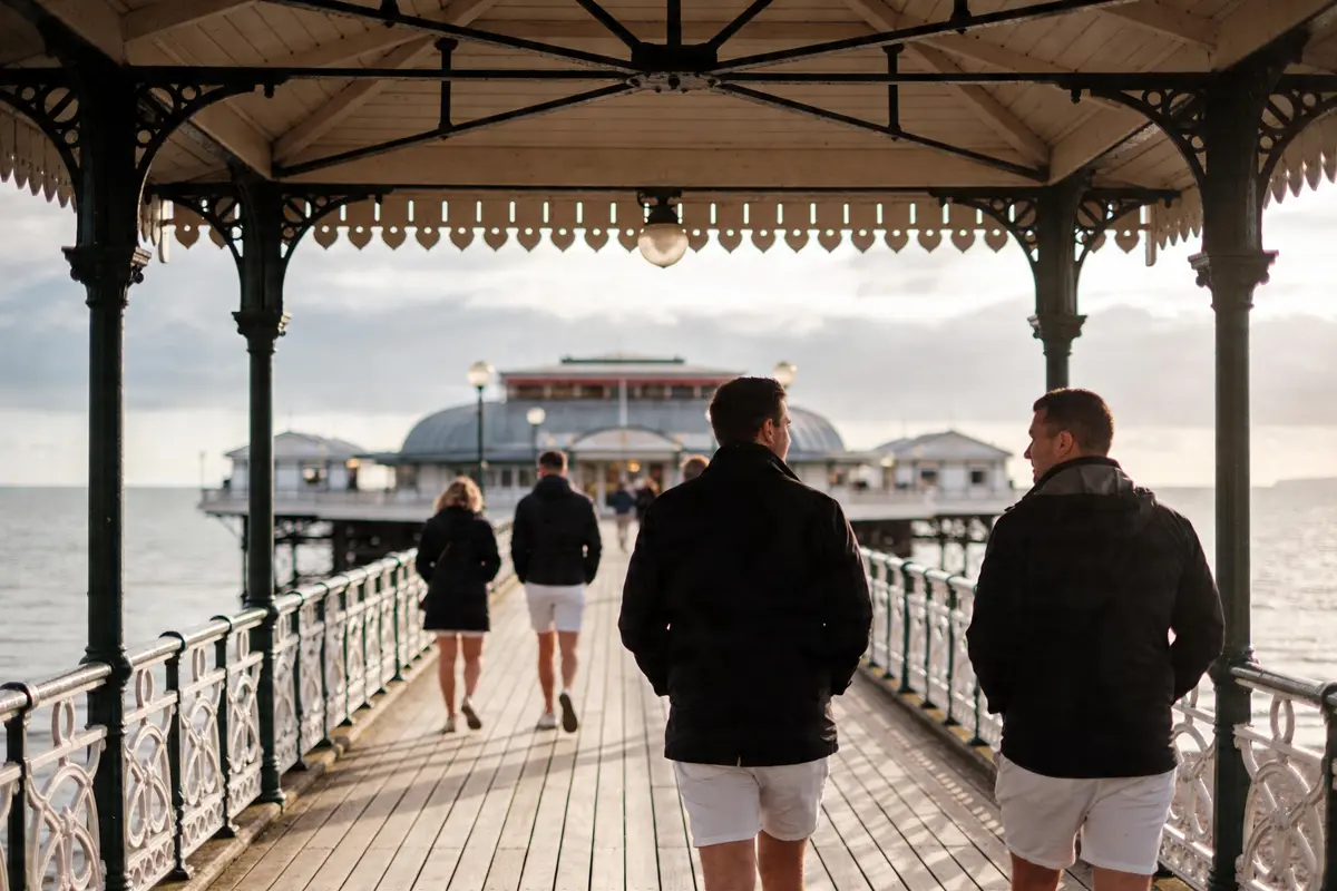 Des personnes marchent sur une jetée en bois, sous un abri, vers un pavillon près de la mer.