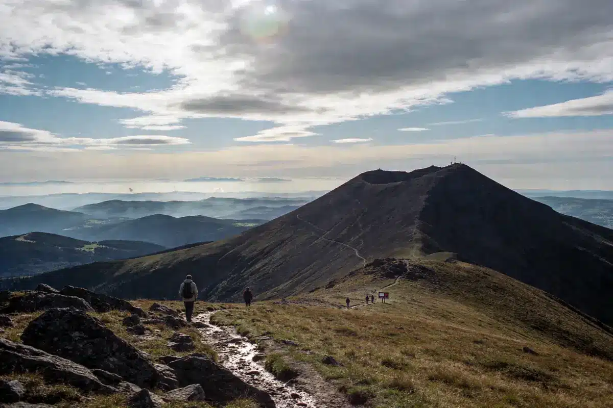 Randonnée en montagne avec sentier en pente, ciel nuageux, randonneurs, paysage de collines et vallée en arrière-plan.