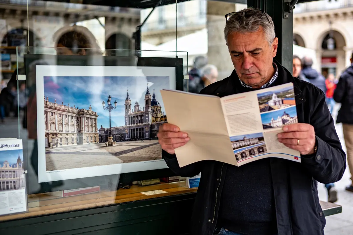Homme lisant une brochure devant une vitrine avec photo d'un palais historique. Passants en arrière-plan.