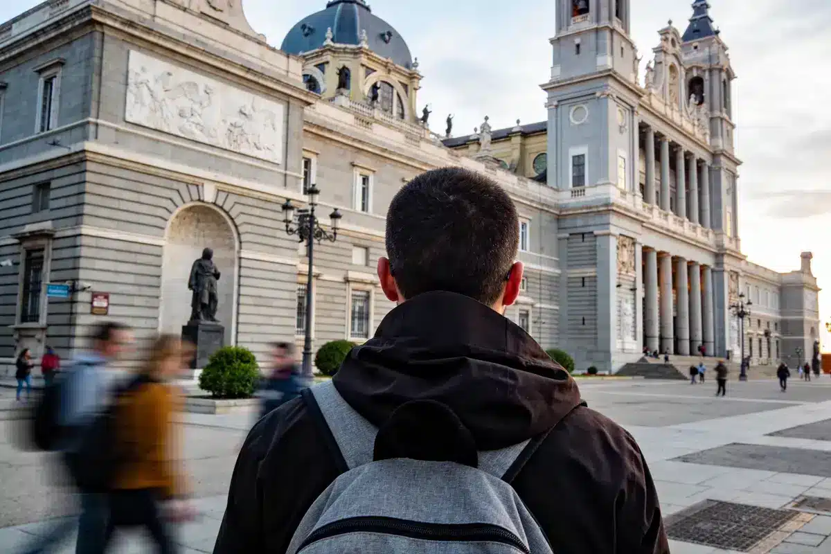 Un homme avec un sac à dos regarde un bâtiment historique en pierre, des passants flous autour.