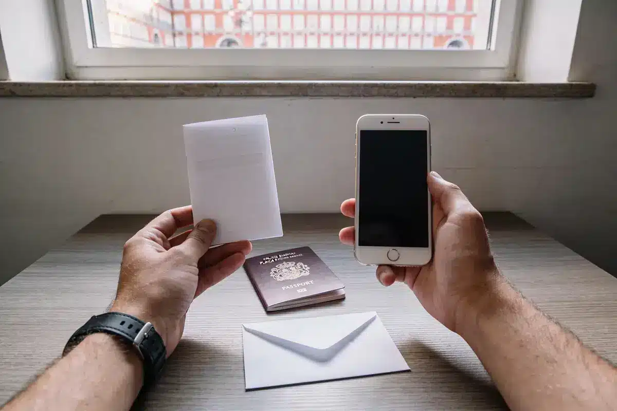 Deux mains tiennent un téléphone et un document, sur une table avec passeport et enveloppe.