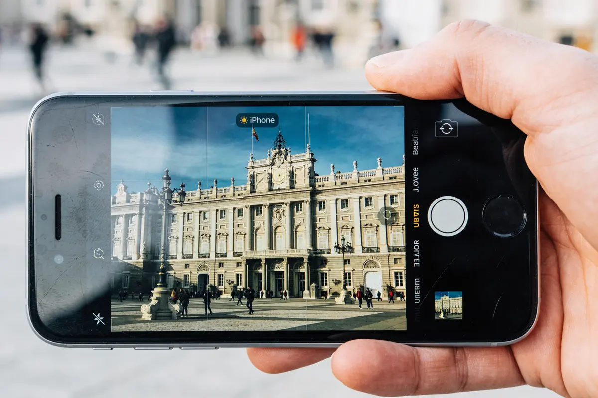 Smartphone avec écran fissuré affichant photo d'un palais. Café et croissant en arrière-plan sur table.