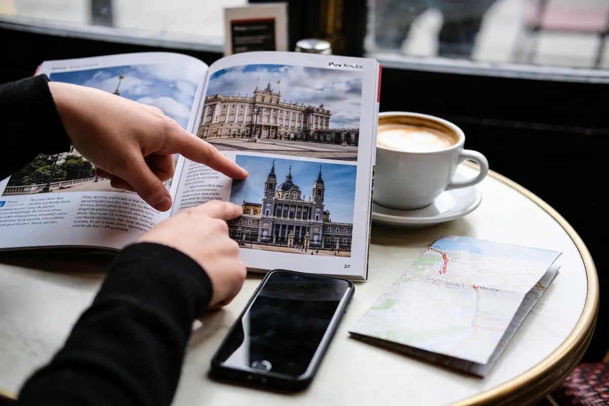 Deux mains pointent un livre illustré sur une table avec téléphone, carte et café.
