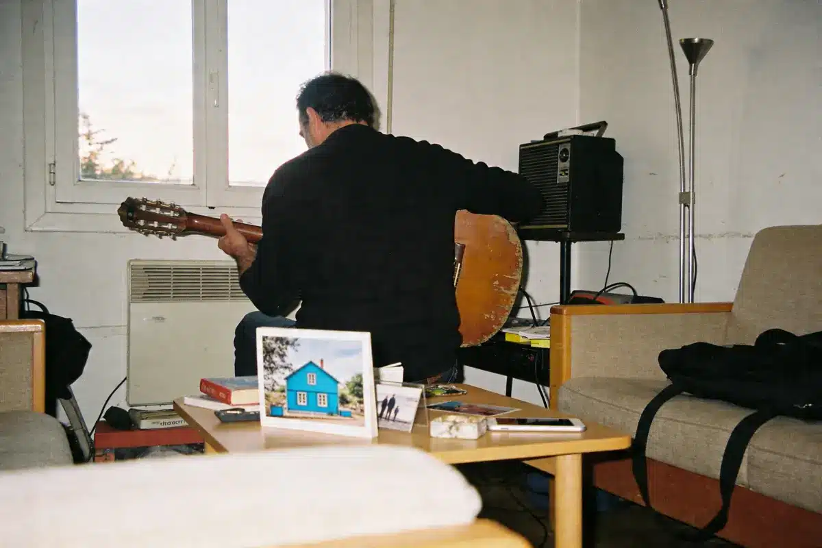 Homme jouant de la guitare dans un salon, table basse avec livres et cartes postales.