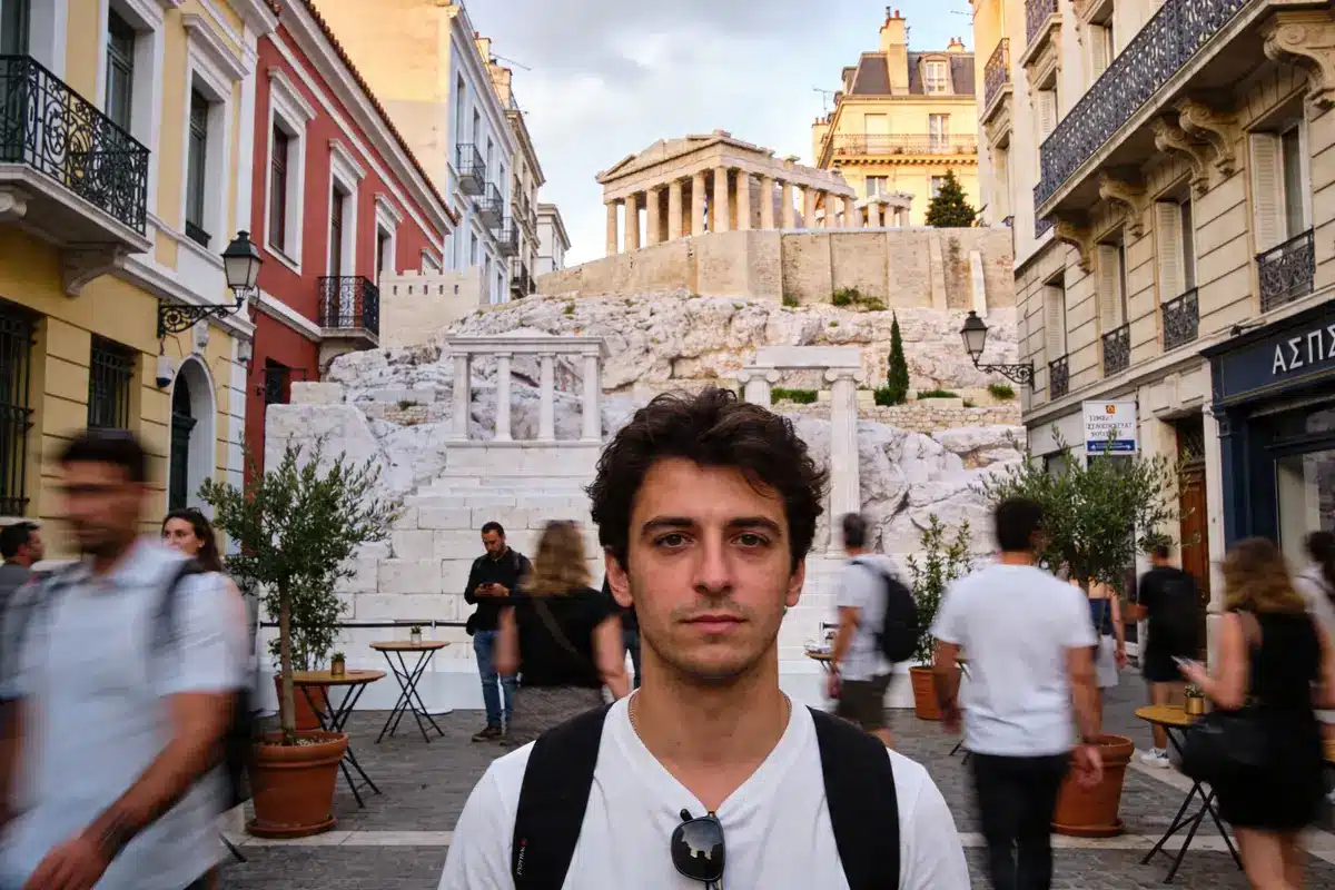 Homme debout dans une rue animée, bâtiments colorés, escaliers et temple en arrière-plan.