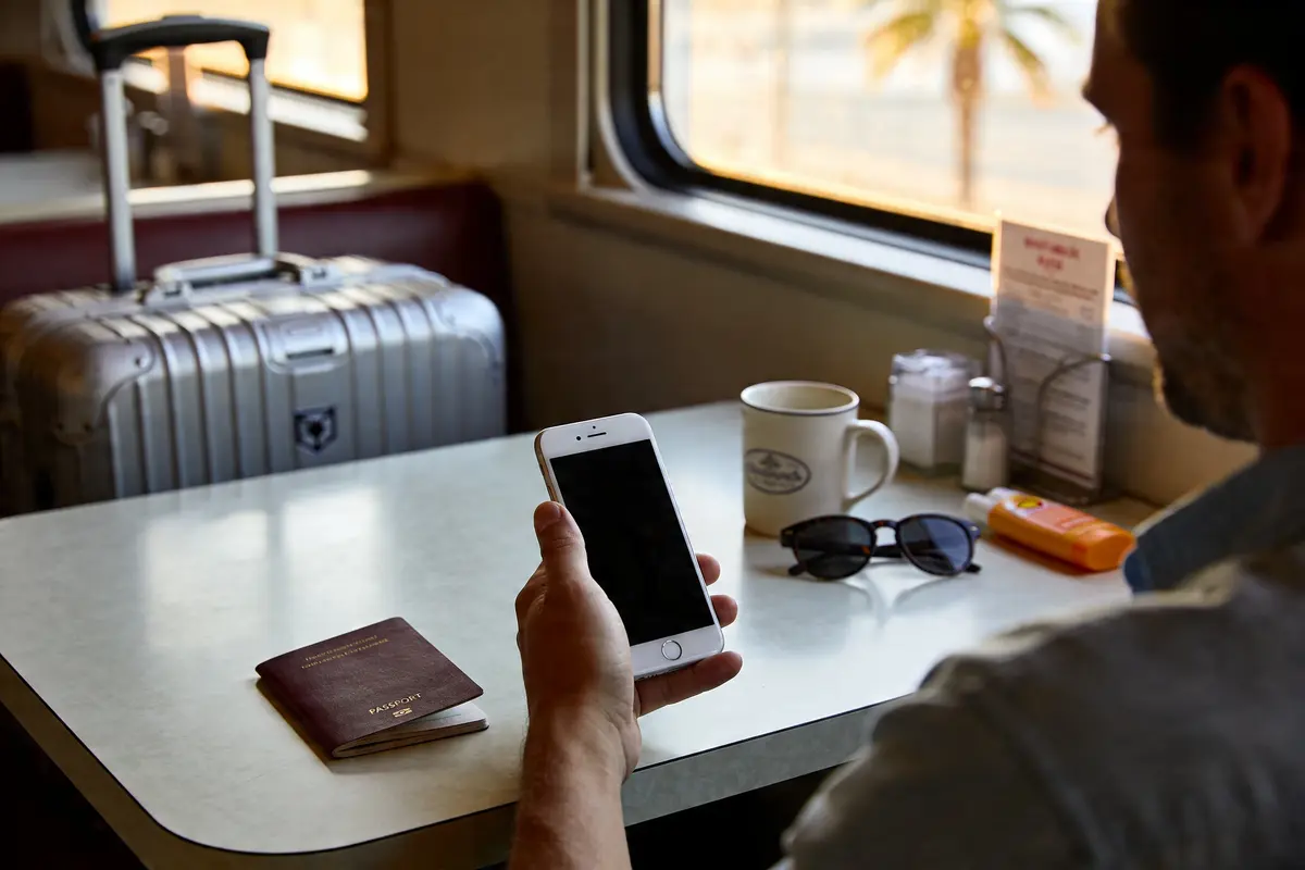 Homme tenant smartphone, assis près d'une table avec passeport, lunettes de soleil, tasse et valise en arrière-plan.