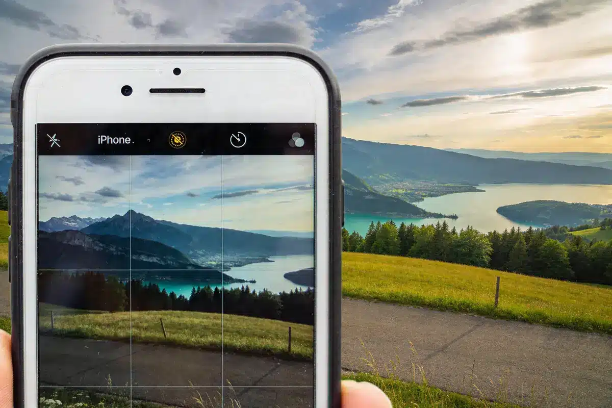 Téléphone prenant photo de paysage montagneux, lac et ciel partiellement nuageux, au coucher du soleil.